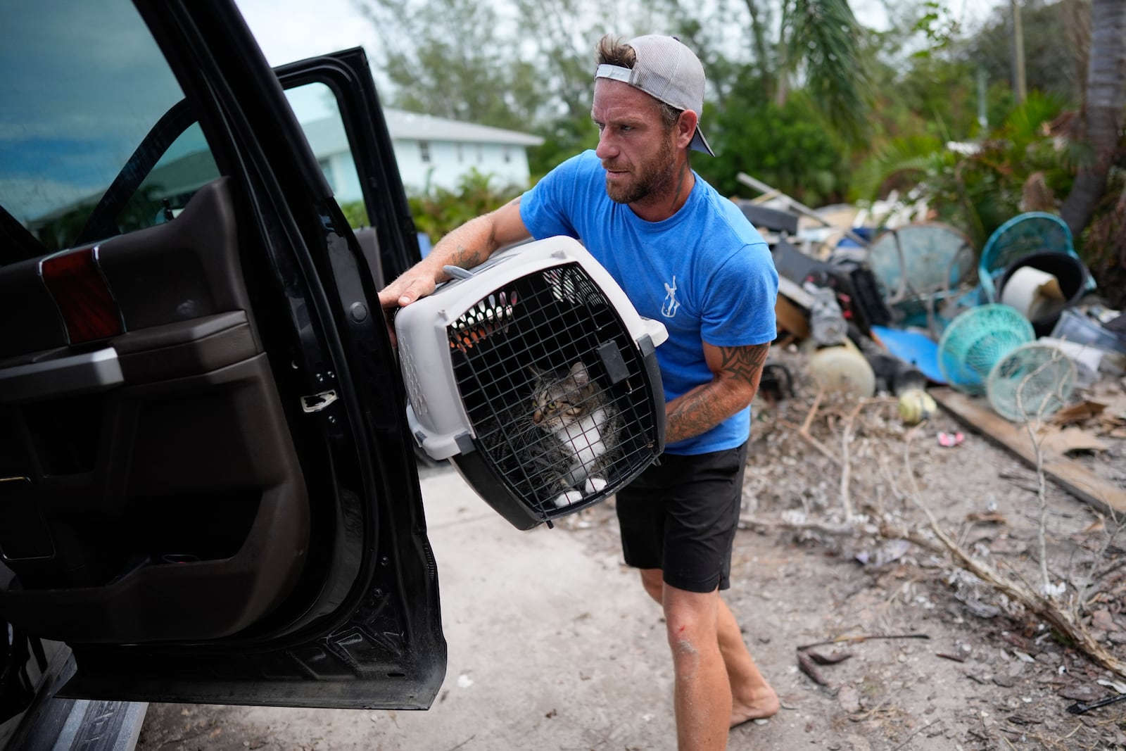 Ted Carlson puts his best friend Evan Purcell's cat McKenzie into a pick-up truck as the pair recover her along with other important items from Purcell's home ahead of the arrival of Hurricane Milton, as debris from Hurricane Helene damage to Purcell's garage level still sits alongside the driveway, in Holmes Beach on Anna Maria Island, Fla., Tuesday, Oct. 8, 2024. "This place couldn't handle Helene," said Carlson, predicting. "It's all going to be gone." (AP Photo/Rebecca Blackwell)