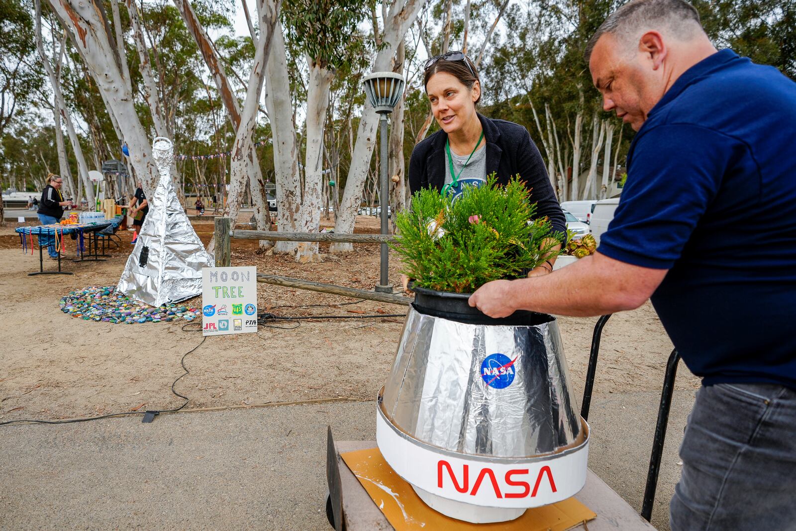 Santiago parent volunteer Stacie Aguesse, left, delivers a small Giant Sequoia tree from NASA's Artemis I Mission's tree seeds that traveled around the moon twice, as NASA scientists, JPL engineers, US Forest Service representatives, and teachers join Santiago STEAM Magnet Elementary School students at a ceremony to plant it after the school was honored in the spring of 2024 to become NASA Moon Tree Stewards in Lake Forest, Calif., on Monday, Oct. 14, 2024. (AP Photo/Damian Dovarganes)