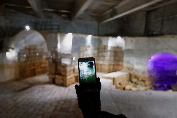 A worker holds up a cell phone displaying a photo of the Ein el-Fijeh reservoir, now seen dry in the background, in the Barada Valley, Syria, Thursday, May 8, 2025. The Ein el-Fijeh spring, the main water source for Damascus, has nearly dried up, forcing many residents in the capital and its suburbs to rely on water delivered by tanker trucks that fill from local wells. (AP Photo/Omar Sanadiki)