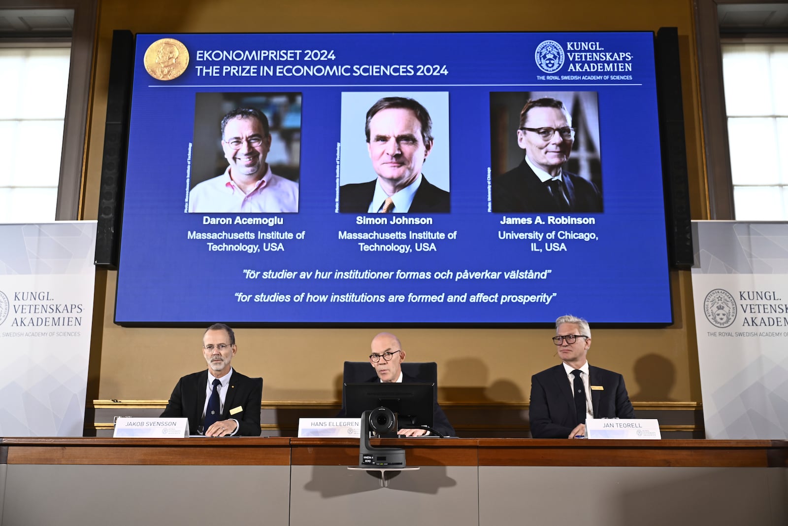 Academy of Sciences permanent secretary Hans Ellegren, center, Jakob Svensson, left, and Jan Teorell, of the Nobel assembly announce the Nobel memorial prize in economics winners, Daron Acemoglu, Simon Johnson and James A Robinson, seen on screen, during a press meeting at the Royal Swedish Academy of Sciences in Stockholm, Sweden, Monday Oct. 14, 2024. (Christine Olsson/TT News Agency via AP)