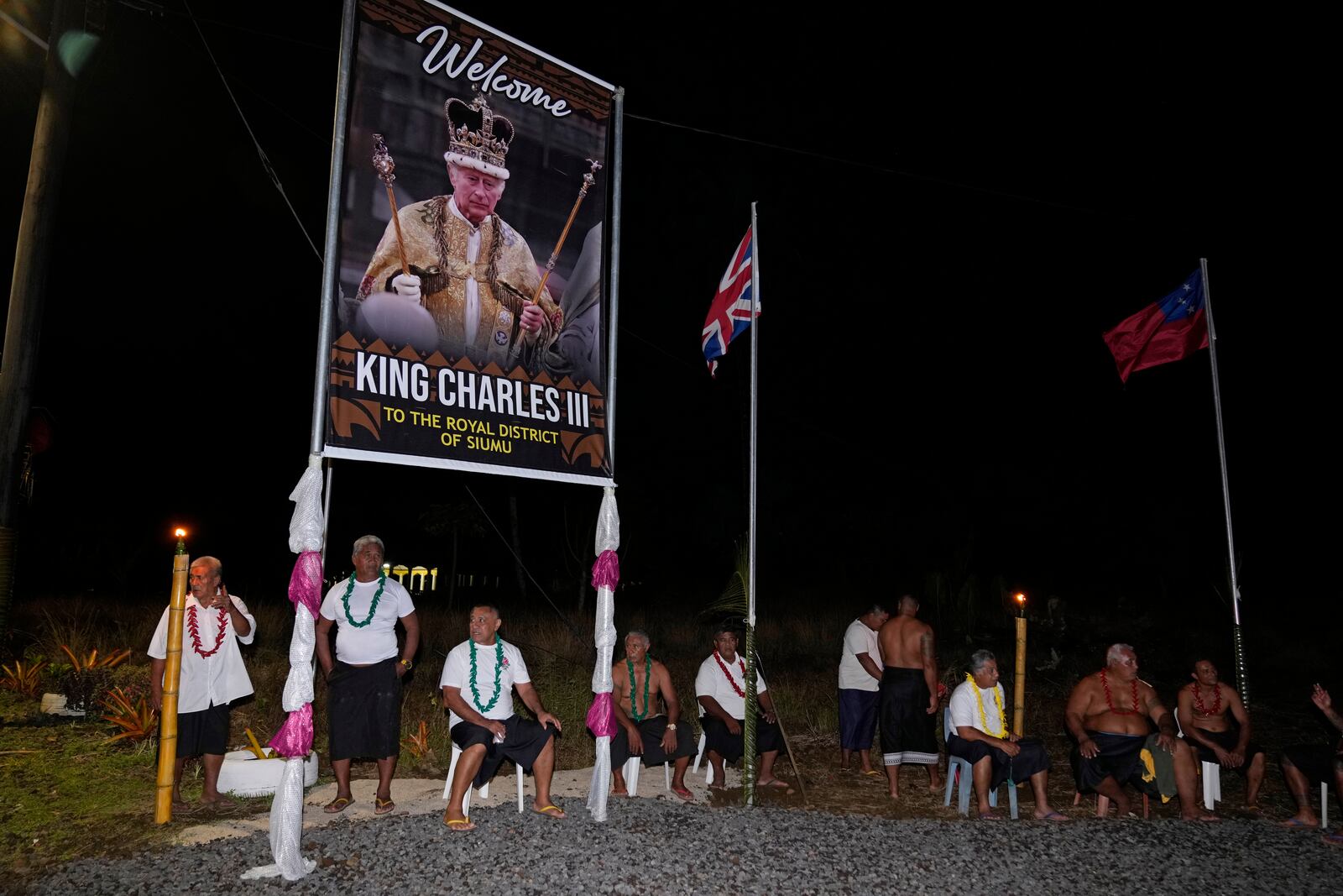 People sit under a portrait of Britain's King Charles III as they wait for his arrival and Queen Camilla in the village of Siumu, Samoa, on Wednesday, Oct. 23, 2024. (AP Photo/Rick Rycroft)