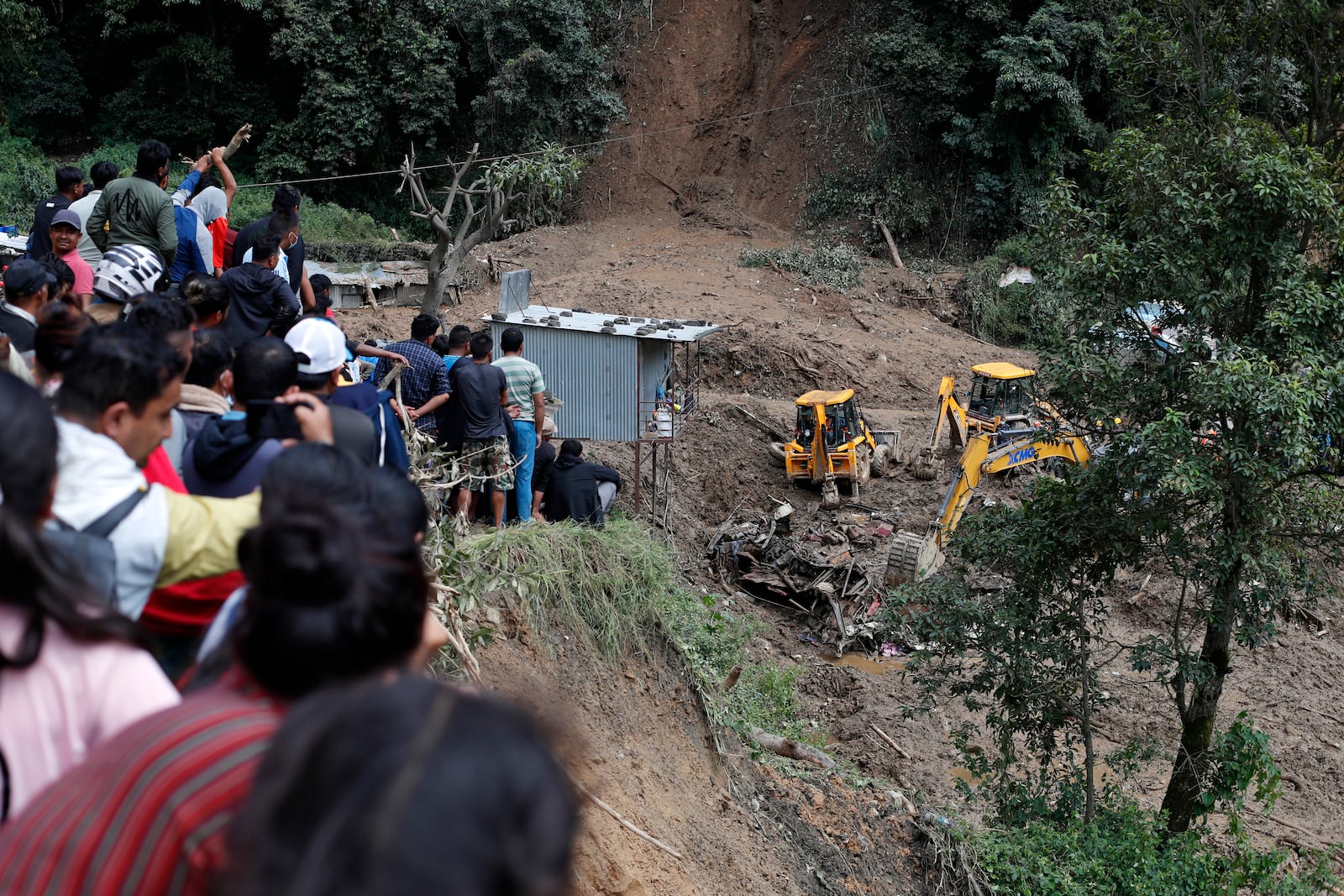 People watch earthmovers removing automobile debris and the dead bodies of victims trapped under a landslide caused by heavy rains in Kathmandu, Nepal, Sunday, Sept. 29, 2024. (AP Photo/Sujan Gurung)