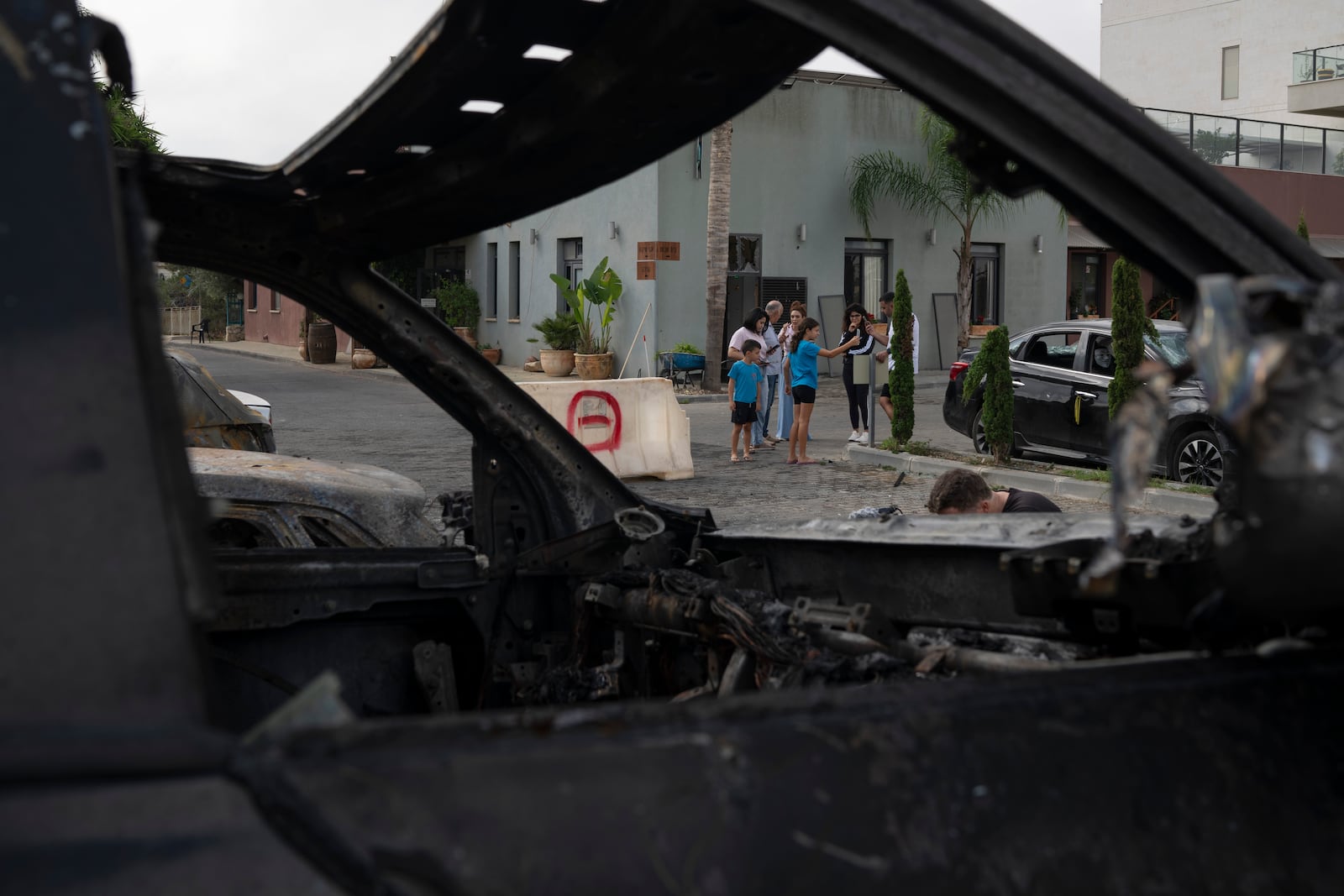 People stand next to burnt and damaged cars after a rocket, launched from Lebanon, hit an area in Kfar Vradim, northern Israel, Monday, Oct. 7, 2024. (AP Photo/Leo Correa)