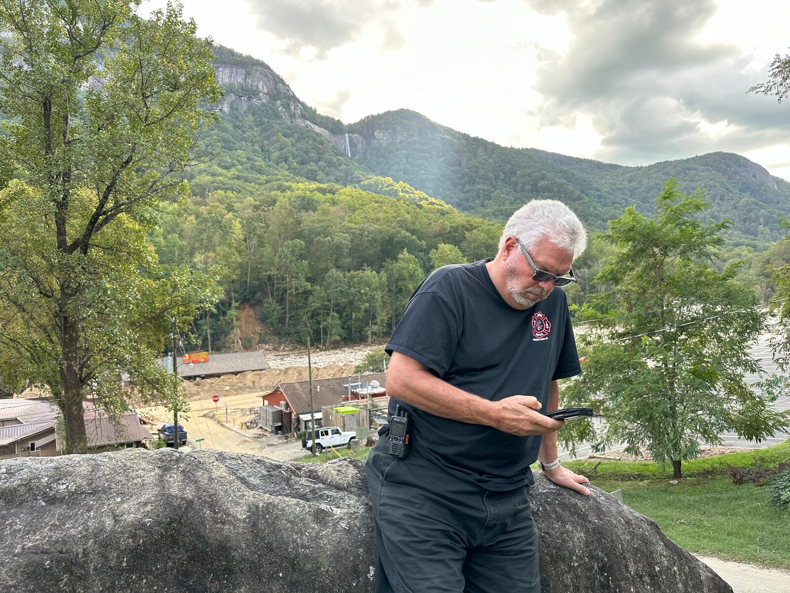 Volunteer firefighter Ric Thurlby checks messages as Hickory Nut Falls flows above and the ruined town of Chimney Rock Village, N.C., lies below on Wednesday, Oct. 2, 2024. (AP Photo/Allen G. Breed)