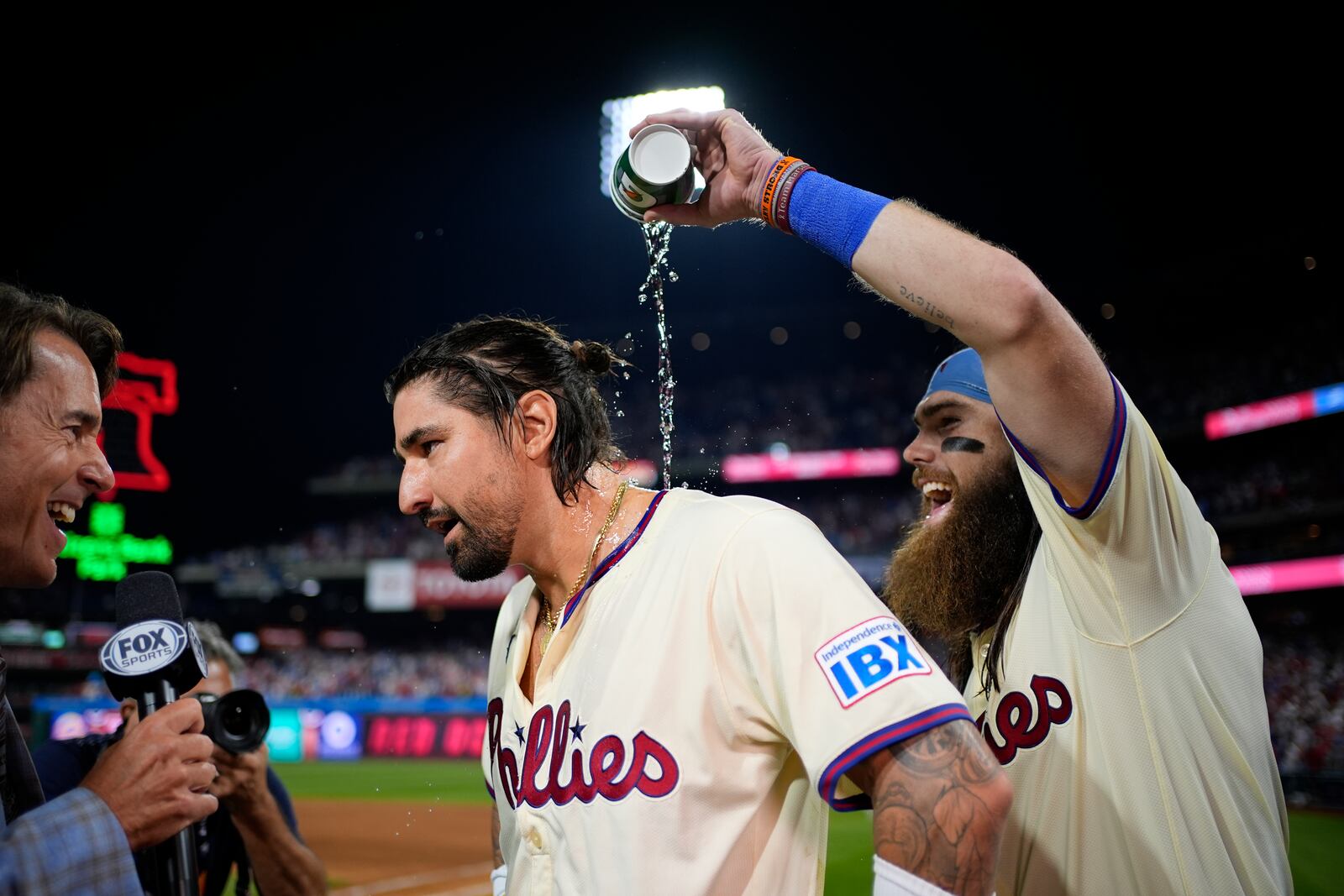 Philadelphia Phillies' Brandon Marsh, right, pours water over Nick Castellanos, center while he is interviewed after the Phillies won Game 2 of a baseball NL Division Series against the New York Mets, Sunday, Oct. 6, 2024, in Philadelphia. (AP Photo/Matt Slocum)