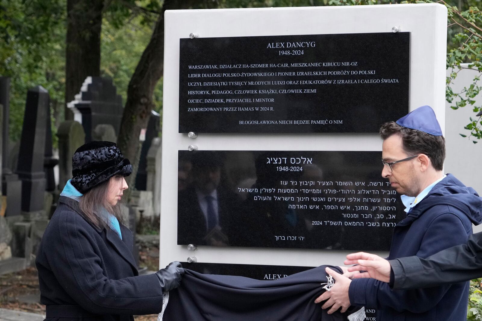 Yuval Danzig, right, the son of a Polish-Israeli man kidnapped and killed by Hamas, helps to unveil a plaque honoring his father, Alex Dancyg, in the Jewish cemetery on the one-year anniversary of the Hamas attack, in Warsaw, Poland, on Monday, Oct. 7, 2024. (AP Photo/Czarek Sokolowski)