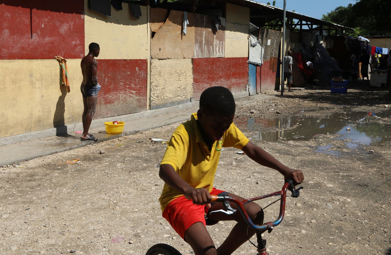 A youth cycles past a man taking a bucket bath at a school where people displaced by gang violence are living in Port-au-Prince, Haiti, Friday, Sept. 20, 2024. (AP Photo/Odelyn Joseph)