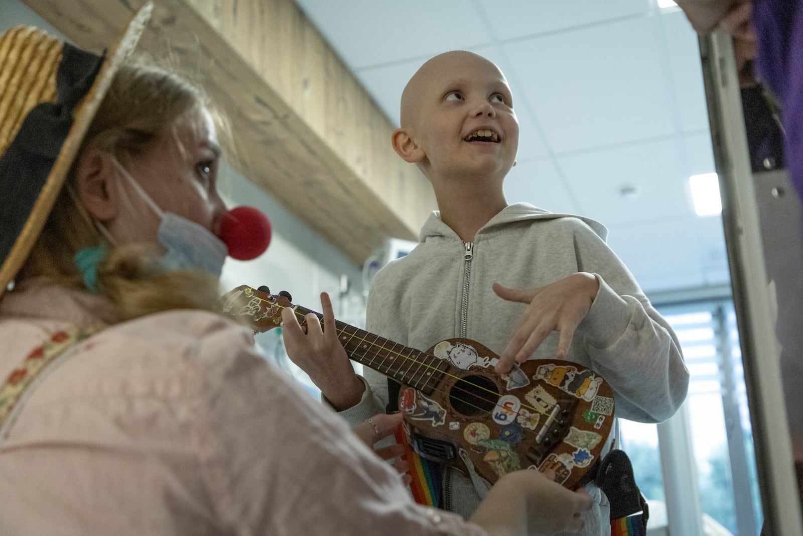 Tetiana Nosova, who goes by the clown name of Zhuzha, a volunteer from the "Bureau of Smiles and Support" watches as Kira Vertetska, 8, plays a ukulele at Okhmatdyt children's hospital in Kyiv, Ukraine, Thursday Sept. 19, 2024. (AP Photo/Anton Shtuka)