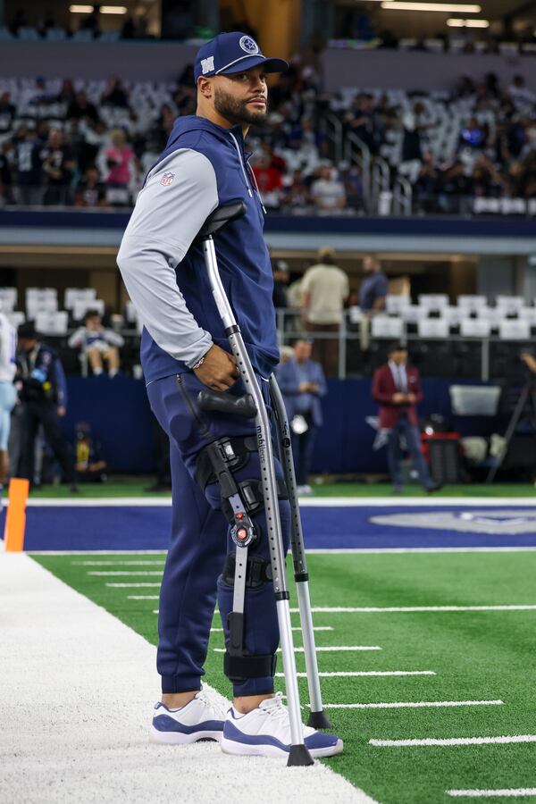 Dallas Cowboys quarterback Dak Prescott stands with crutches on the sidelines as teammates work out prior to an NFL football game against the Houston Texans, Monday, Nov. 18, 2024, in Arlington. (AP Photo/Gareth Patterson)