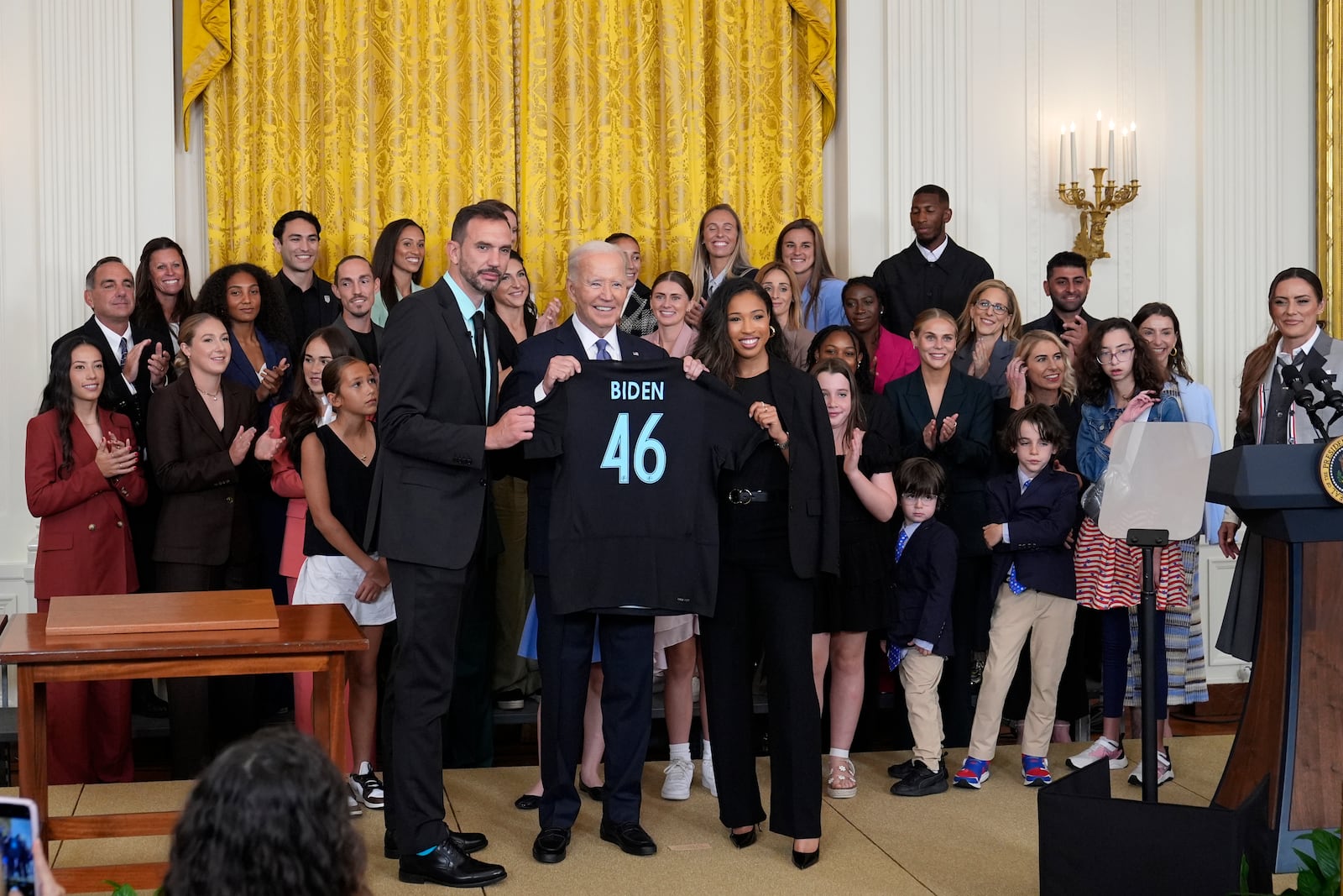 President Joe Biden, center, holds a team jersey from NJ/NY Gotham FC that was presented to him by head coach Juan Carlos Amoros, left, and teammate Midge Purse, right, during an event in the East Room of the White House in Washington, Monday, Sept. 23, 2024, to welcome the NJ/NY Gotham FC and celebrate their 2023 NWSL championship. (AP Photo/Susan Walsh)